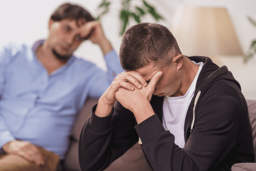 Man sitting on a couch with his head in his hands with another man in the background looking at him